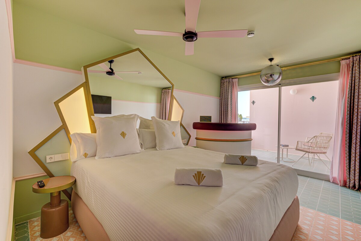 A hotel room with a large bed, a ceiling fan, a bathroom with a round mirror, and a view of the outside.