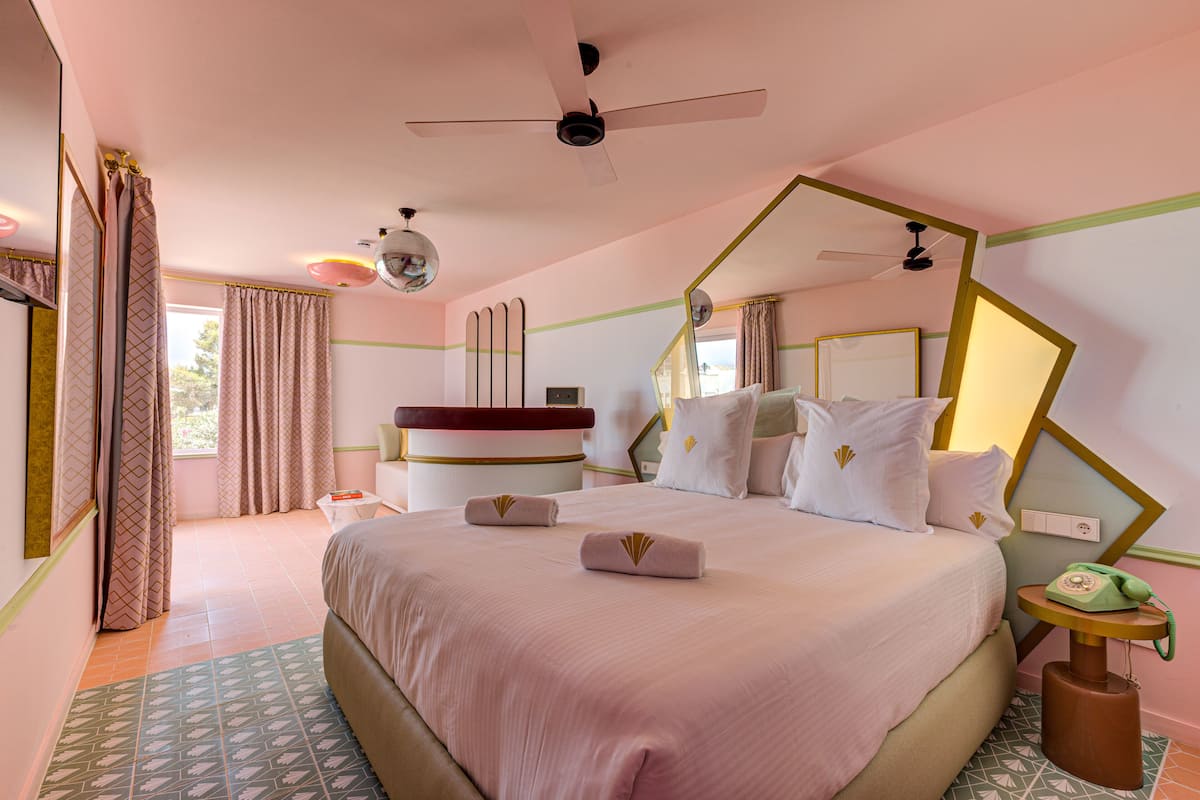 Quarto de hotel com uma cama grande, um ventilador de teto, uma mesinha de cabeceira com telefone verde e um espelho na parede.