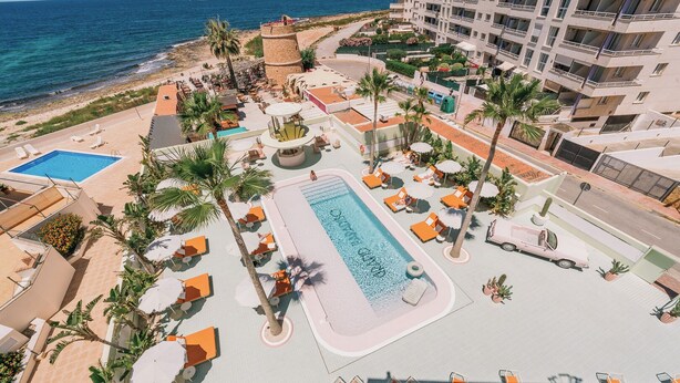 A resort with a central swimming pool, multiple lounge areas with umbrellas, and a view of the ocean.