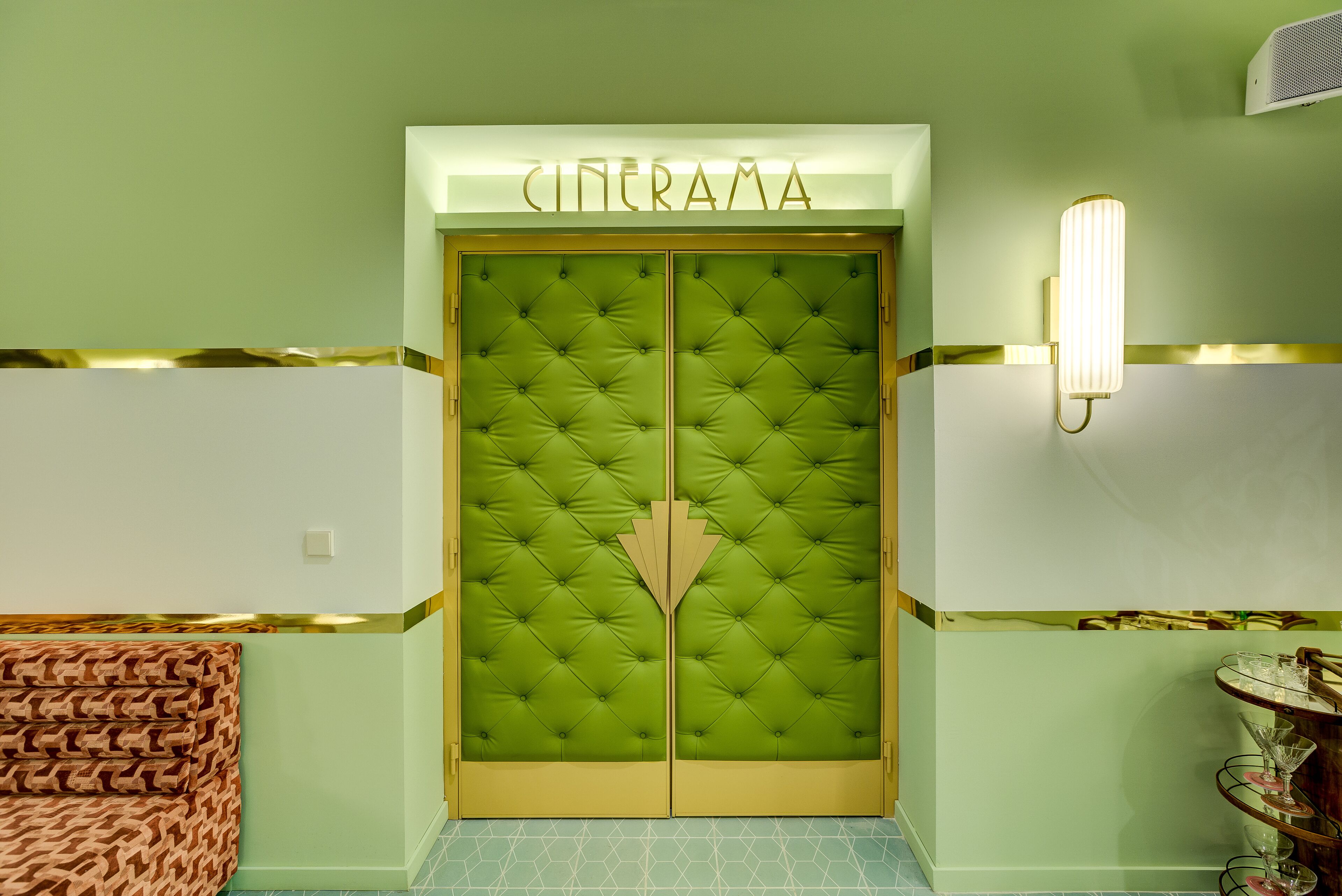 A green door with the word "CINERAMA" above it, a tufted design, and a wall-mounted light fixture to the right.