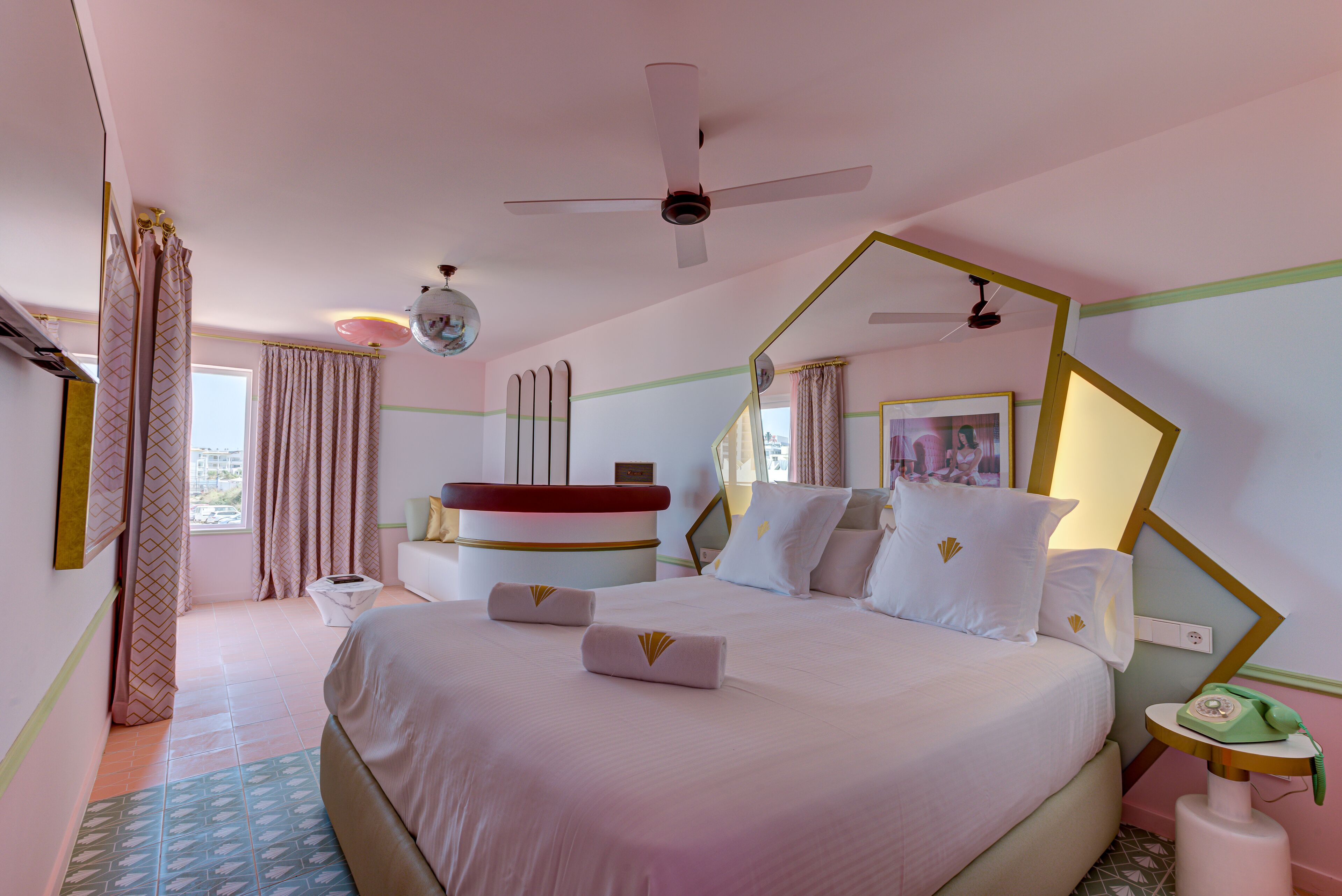 Una habitación de hotel con una cama grande, un ventilador de techo, un escritorio con teléfono y una ventana con cortinas.