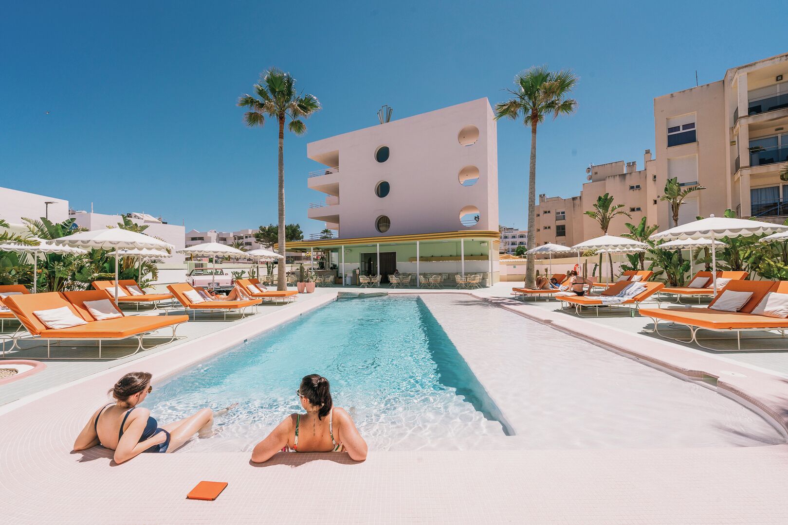 A pool area with lounge chairs and umbrellas, a modern building with circular windows, and palm trees.