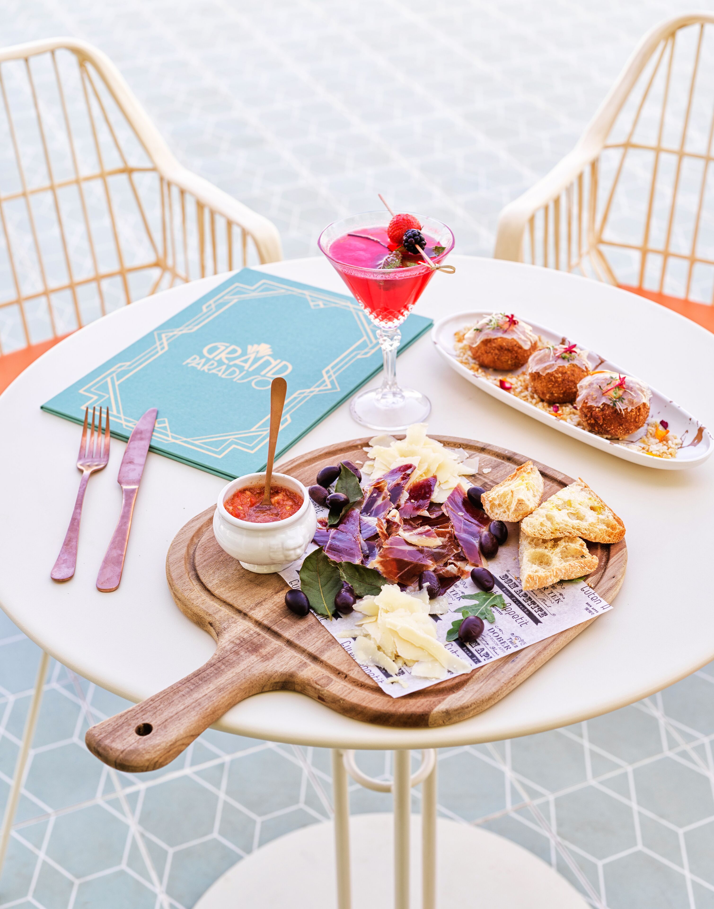 A table set with a blue menu, a martini glass with a berry garnish, a plate of croquettes, a wooden board with cheese and olives, and a small bowl of sauce.