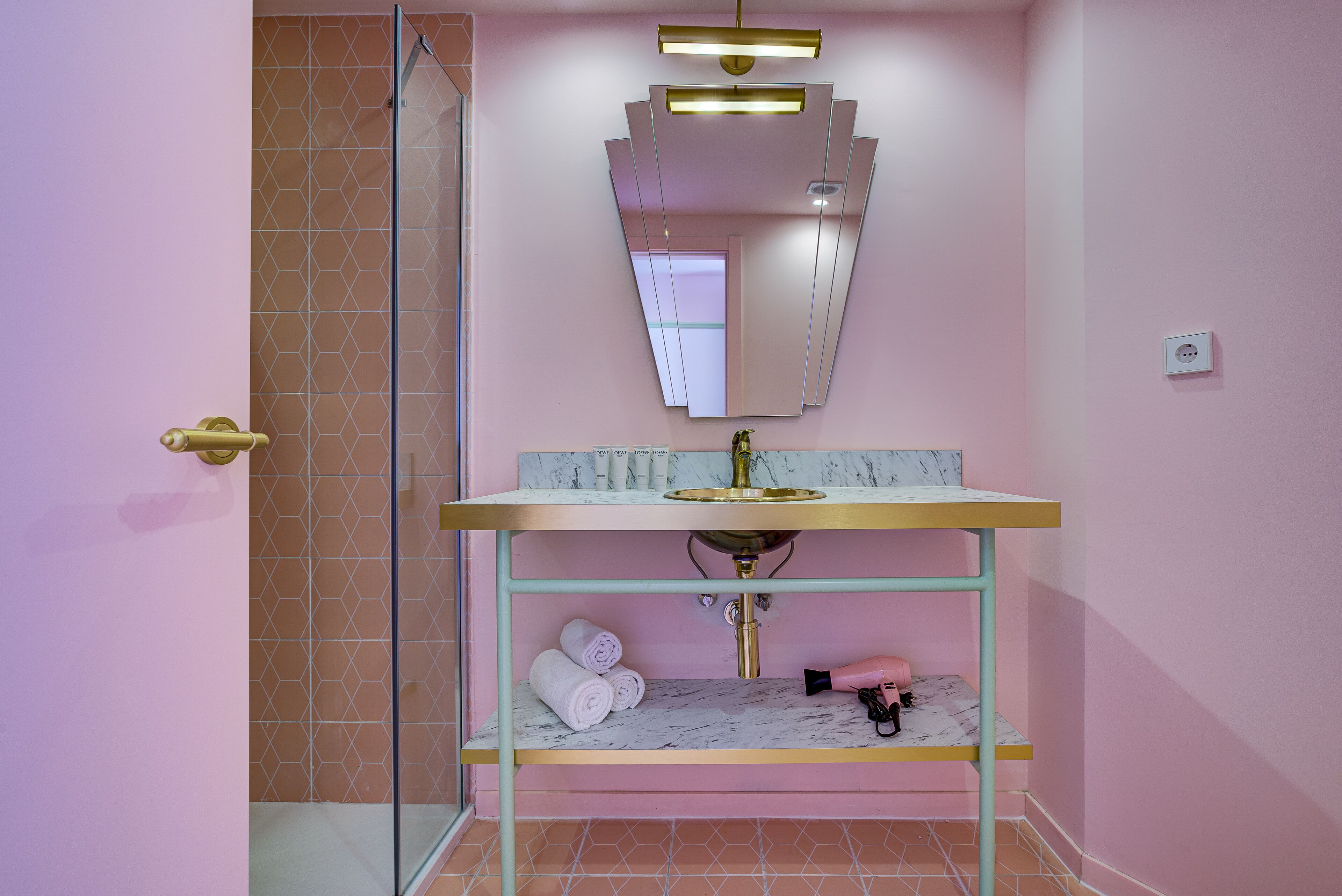 A bathroom with a pink wall, a gold-framed sink, a marble countertop, and a glass shower enclosure.