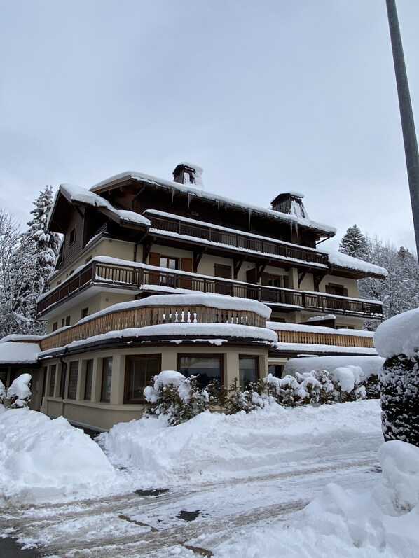 A multi-story building with balconies, surrounded by snow-covered trees and bushes.