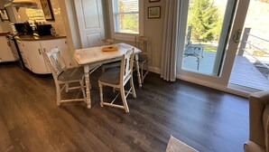 Un coin repas avec une table et des chaises blanches, une cuisine équipée d’une cuisinière et de placards, et une porte-fenêtre coulissante donnant sur une terrasse extérieure.