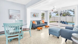 A modern living room with a grey sofa, blue chairs, and a dining table. There is a large window with a view of a swimming pool and outdoor furniture.
