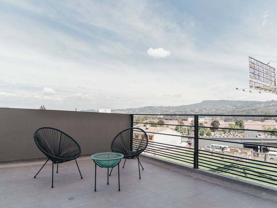 A balcony with two chairs and a small table, overlooking a cityscape with buildings and a distant mountain range.