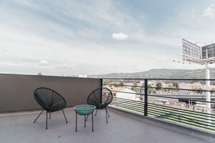 Un balcón con dos sillas y una pequeña mesa, con vistas a un paisaje urbano con edificios y una cadena montañosa distante.