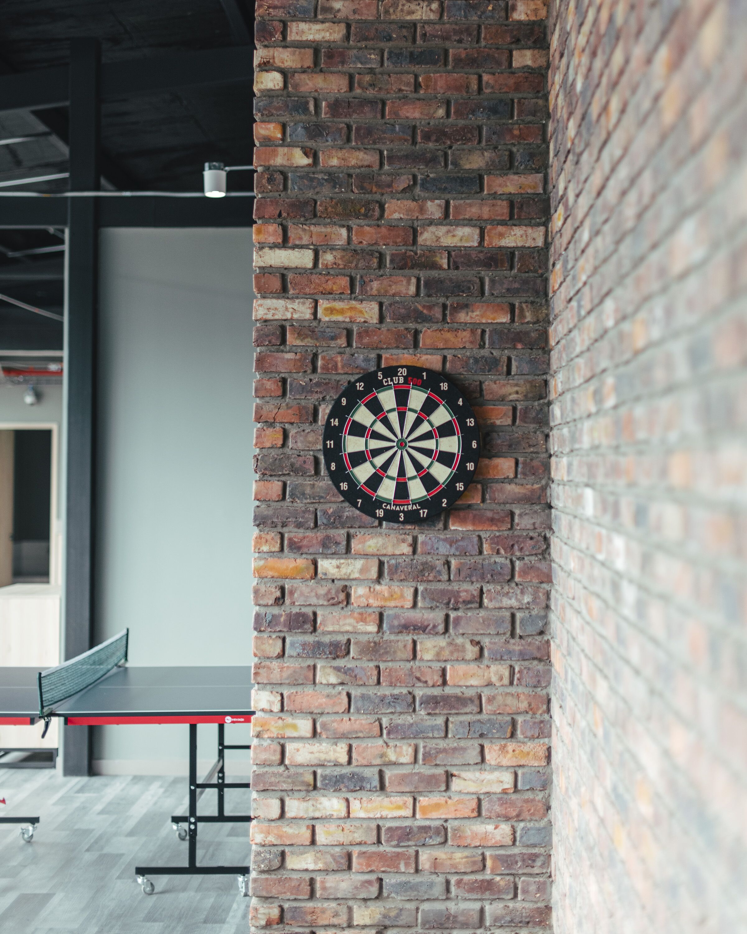 A dartboard mounted on a brick wall with a ping pong table in the background.
