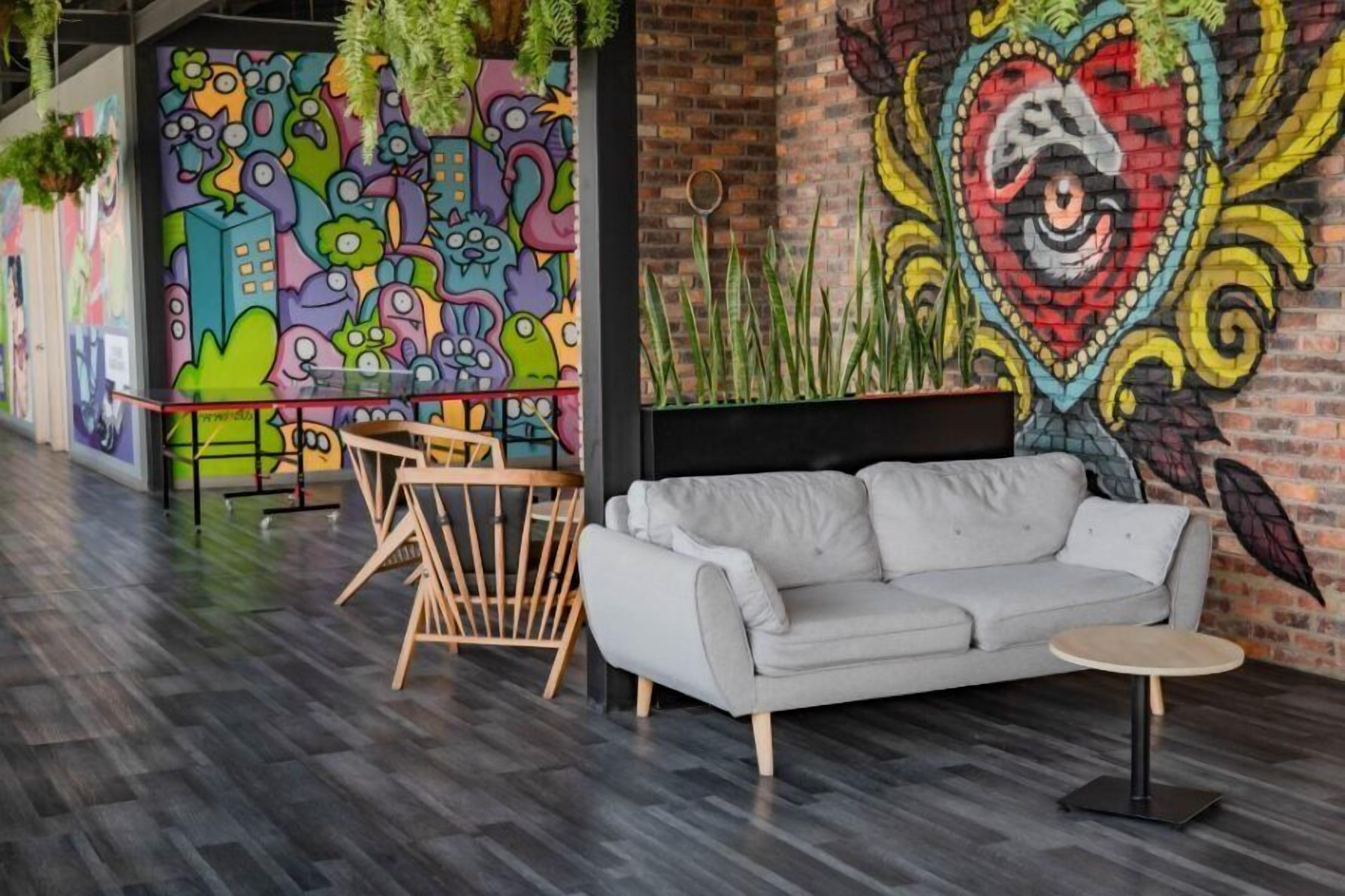 Ein moderner Lounge-Bereich mit grauem Sofa, rundem Tisch und einem Holzstuhl. Die Wände sind mit farbenfrohen Wandgemälden und einem großen, herzförmigen Design verziert.