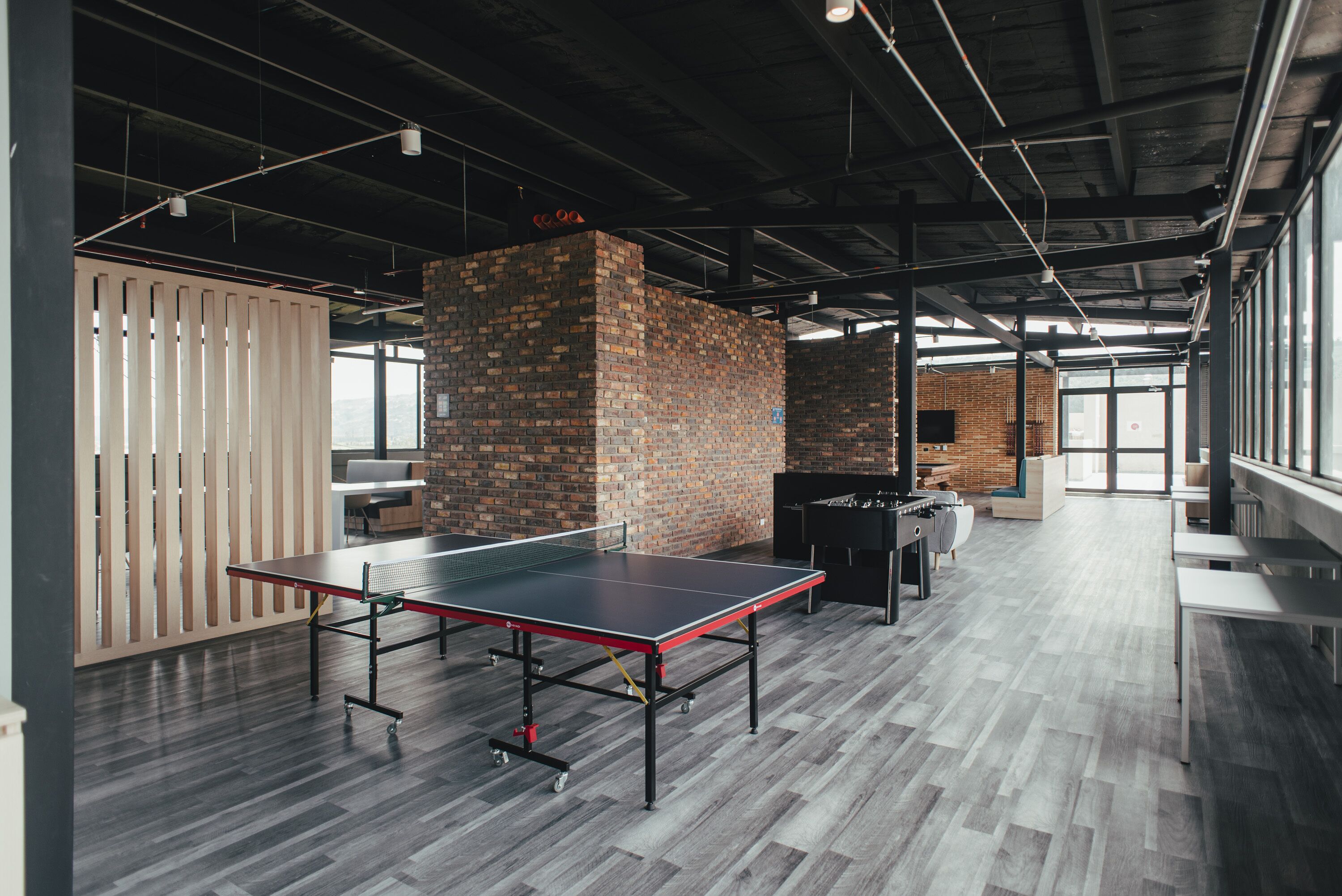Amplio espacio interior con mesa de ping pong, pared de ladrillo y zona de estar con mesas y sillas.