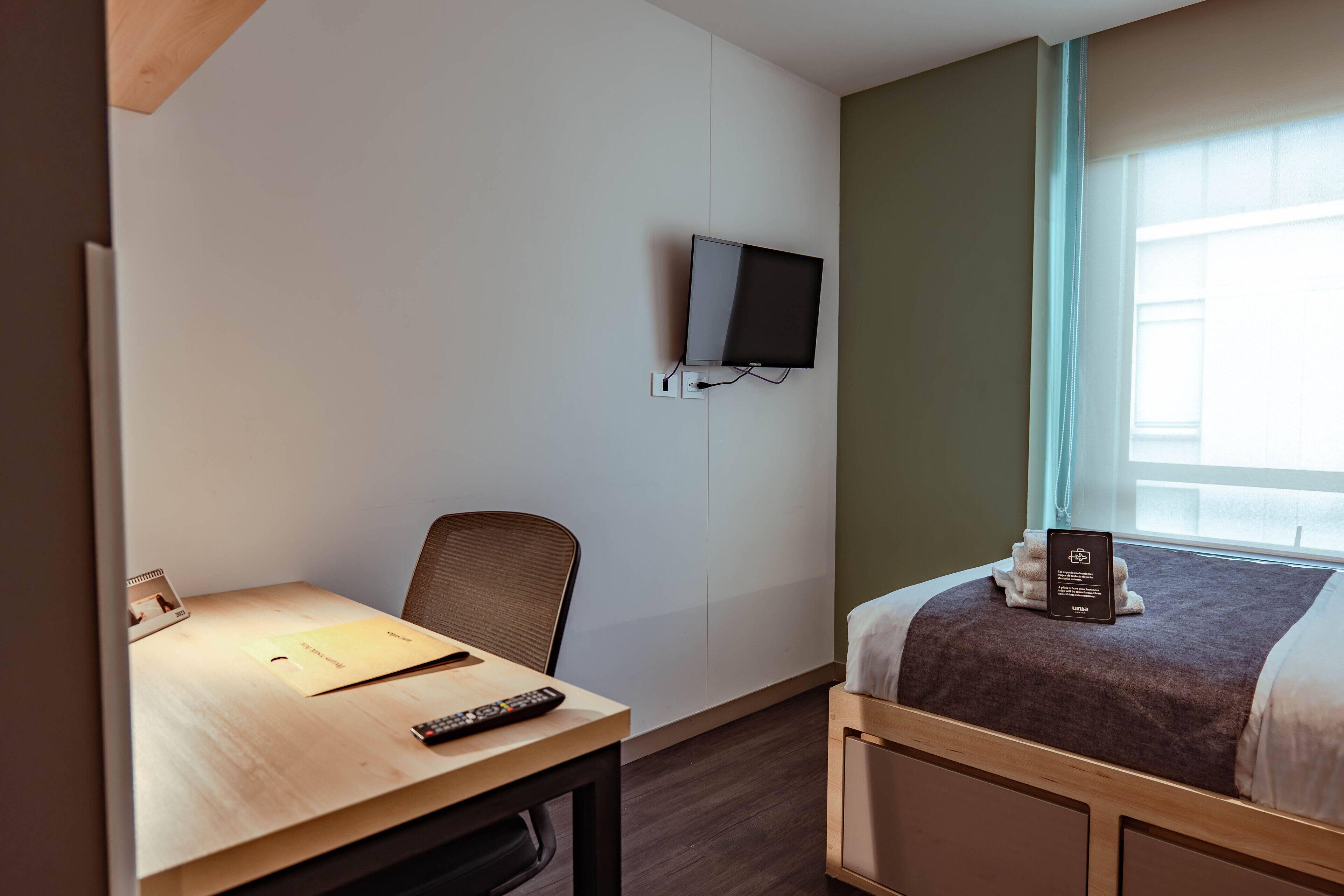 Een hotelkamer met een bed, een bureau met stoel, een aan de muur bevestigde televisie en een raam met gordijnen.