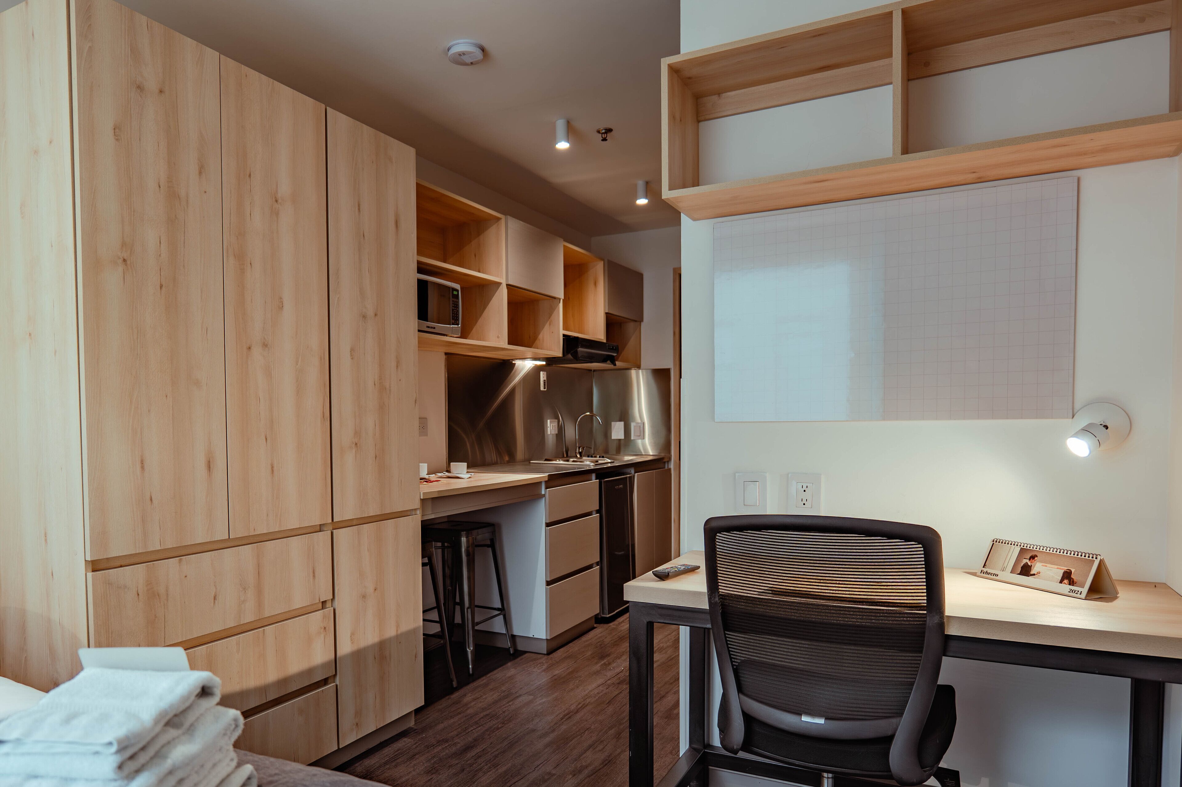 A compact kitchen with wooden cabinets, a stainless steel refrigerator, and a small dining area with a chair and a desk.