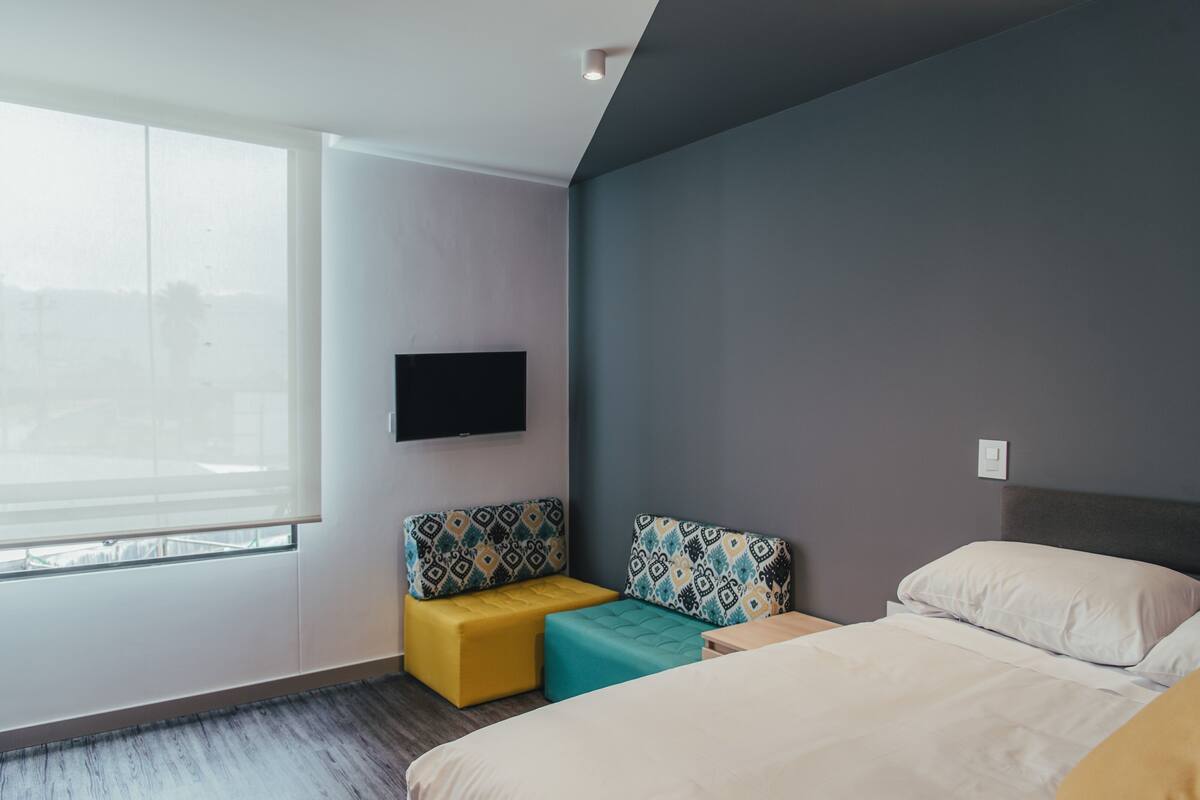 A modern hotel room with a large window, a bed with a headboard, a flat-screen TV mounted on the wall, and two patterned ottomans.