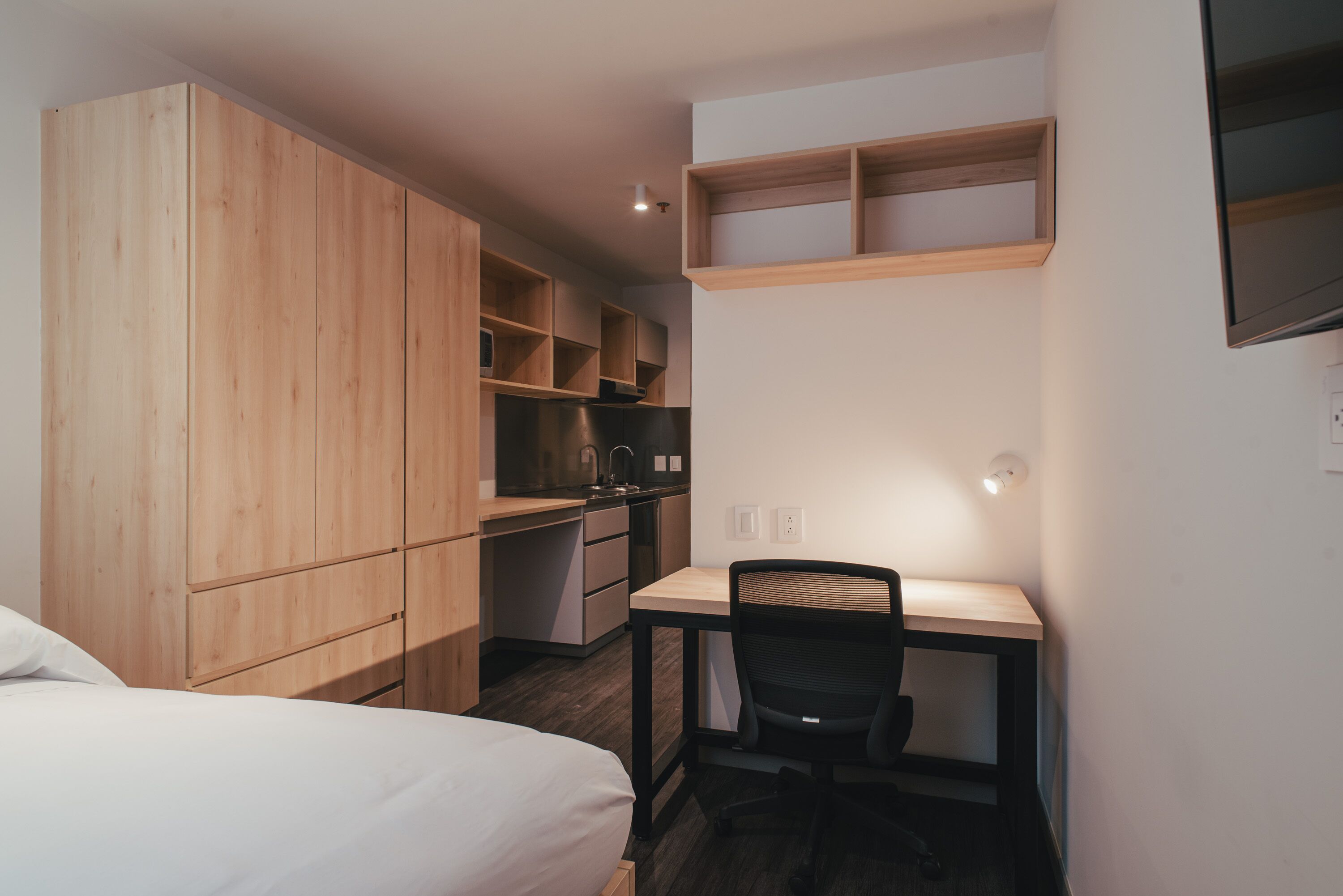 A compact room with a bed, a desk with a chair, a kitchenette, and built-in wooden cabinets.
