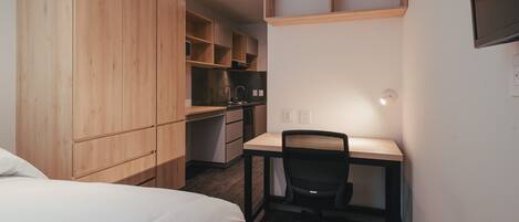 A compact room with a bed, a desk with a chair, a kitchenette, and built-in wooden cabinets.