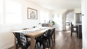 A dining area with a wooden table and black chairs, a kitchen in the background, and a framed picture on the wall.
