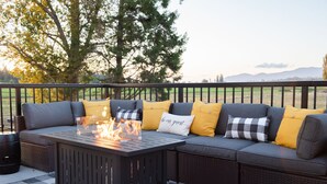 A sectional sofa with yellow and gray cushions, a black metal table with a fire feature, and a checkered rug on an outdoor patio with a view of trees and hills.