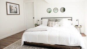 A bedroom with a large bed, a bedside table with a lamp, a framed picture on the wall, and a door.