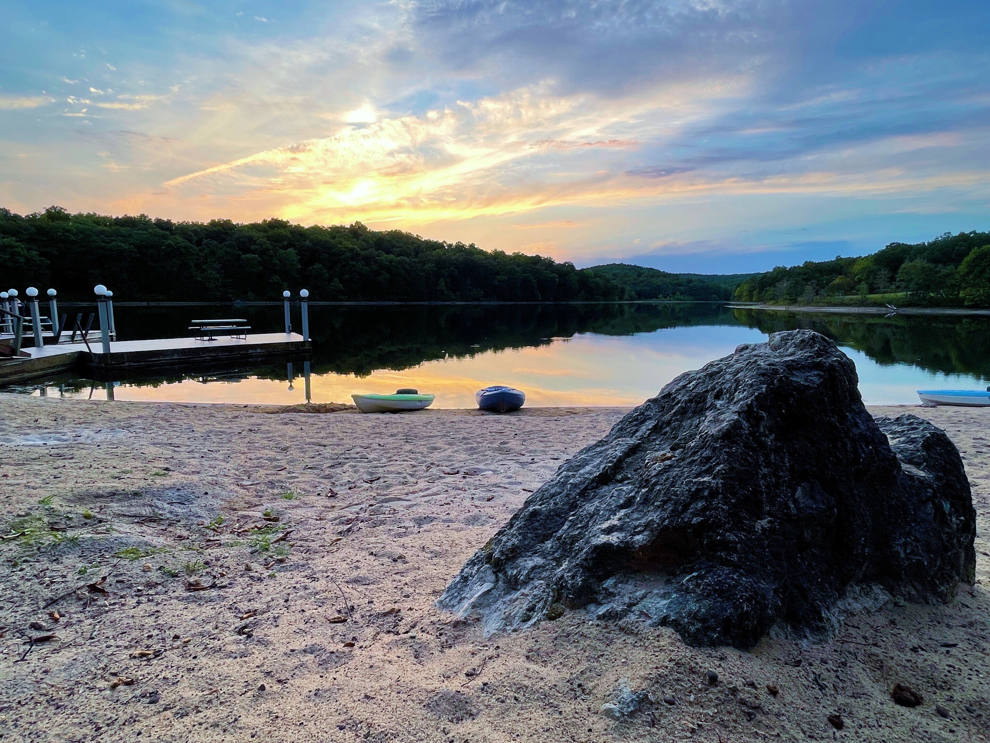 Un lac paisible avec un quai, un grand rocher au premier plan et un coucher de soleil en arrière-plan.