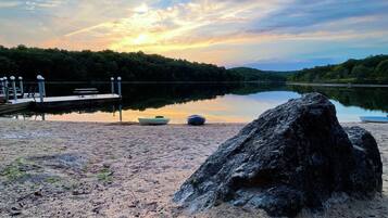 A calm lake with a dock, a large rock in the foreground, and a sunset in the background.