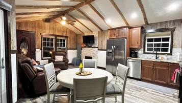 A cozy kitchen with wooden beams, a dining area with chairs, a fireplace, and a refrigerator.