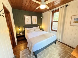 A bedroom with a bed, wooden ceiling, a window with curtains, a wall-mounted light, and framed pictures. - Stonewall Christmas Cottage, Door County, Fire Pit, Coffee Bar - Sleeps 8 (Baileys Harbor)