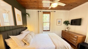 Une chambre à coucher avec un lit, une commode en bois, un ventilateur de plafond et un téléviseur fixé au mur.