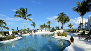 Uma área de piscina com espreguiçadeiras e guarda-sóis, cercada por palmeiras e um céu claro.