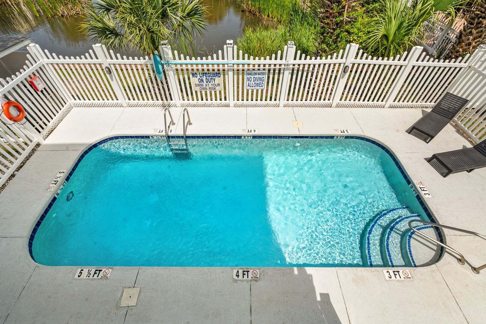 Une piscine rectangulaire entourée d’une clôture blanche, avec des panneaux d’avertissement indiquant une faible profondeur et des chaises longues.