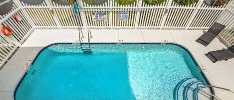 Une piscine rectangulaire entourée d’une clôture blanche, avec des panneaux d’avertissement indiquant une faible profondeur et des chaises longues.