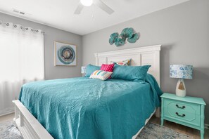 Quarto com colcha azul-esverdeada, cabeceira branca e mesa de cabeceira com abajur.