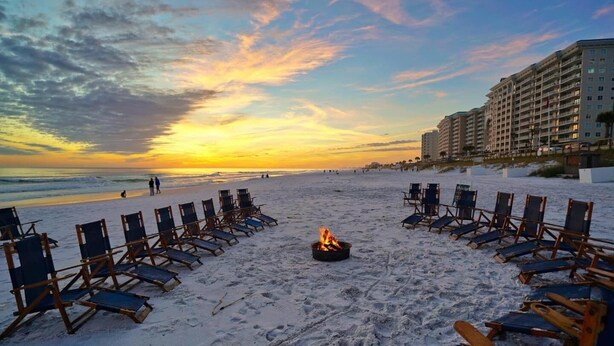 A beach with lounge chairs arranged in a semi-circle around a fire pit, overlooking the ocean and a hotel in the background.