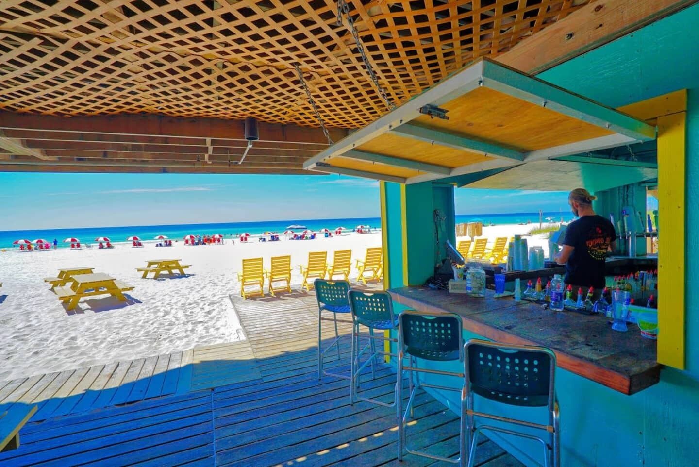 A beach bar with a wooden deck, outdoor seating, and a view of the beach and ocean.