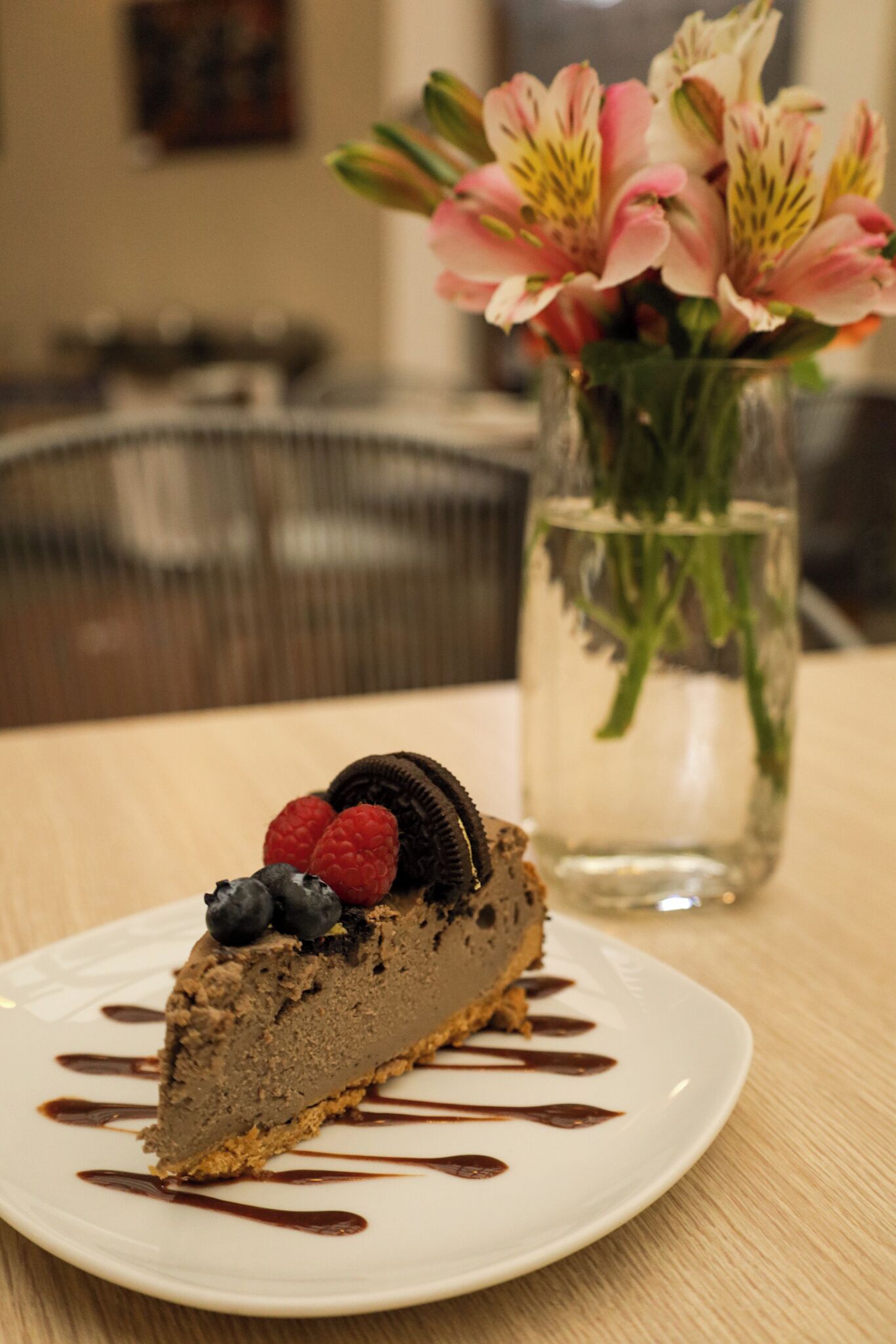 Una porción de pastel de queso con chocolate y frutos rojos sobre un plato, con un jarrón de flores al fondo.