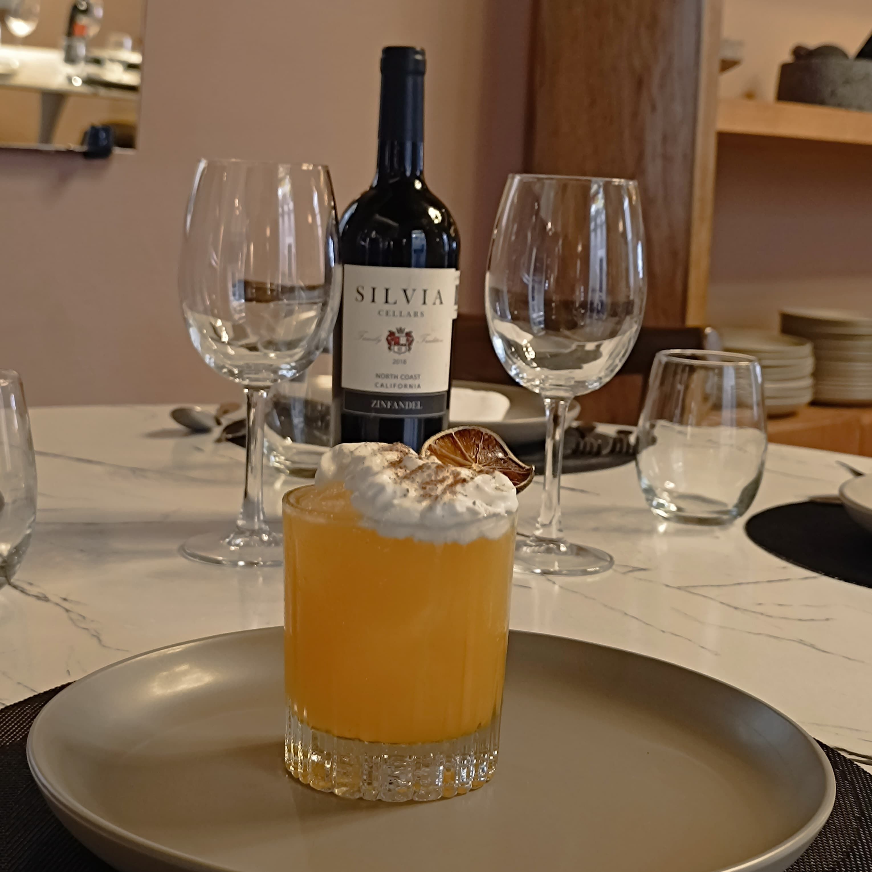 Una mesa servida con un vaso de bebida amarilla adornada con crema batida y una rodaja de fruta, una botella de vino y varias copas de vino vacías.