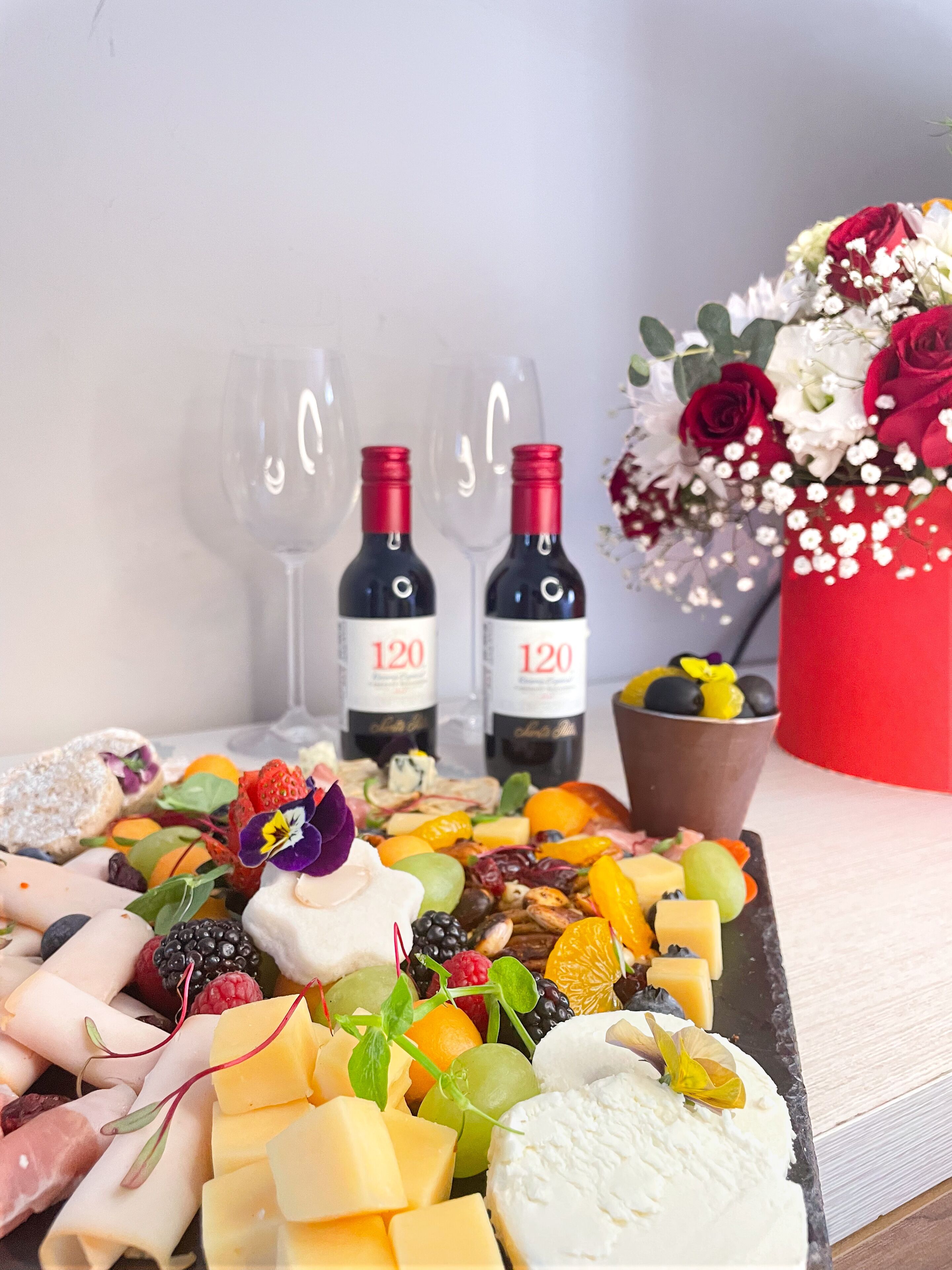 Een charcuterieplank met diverse kazen, fruit en noten, vergezeld van twee flessen wijn en lege wijnglazen.