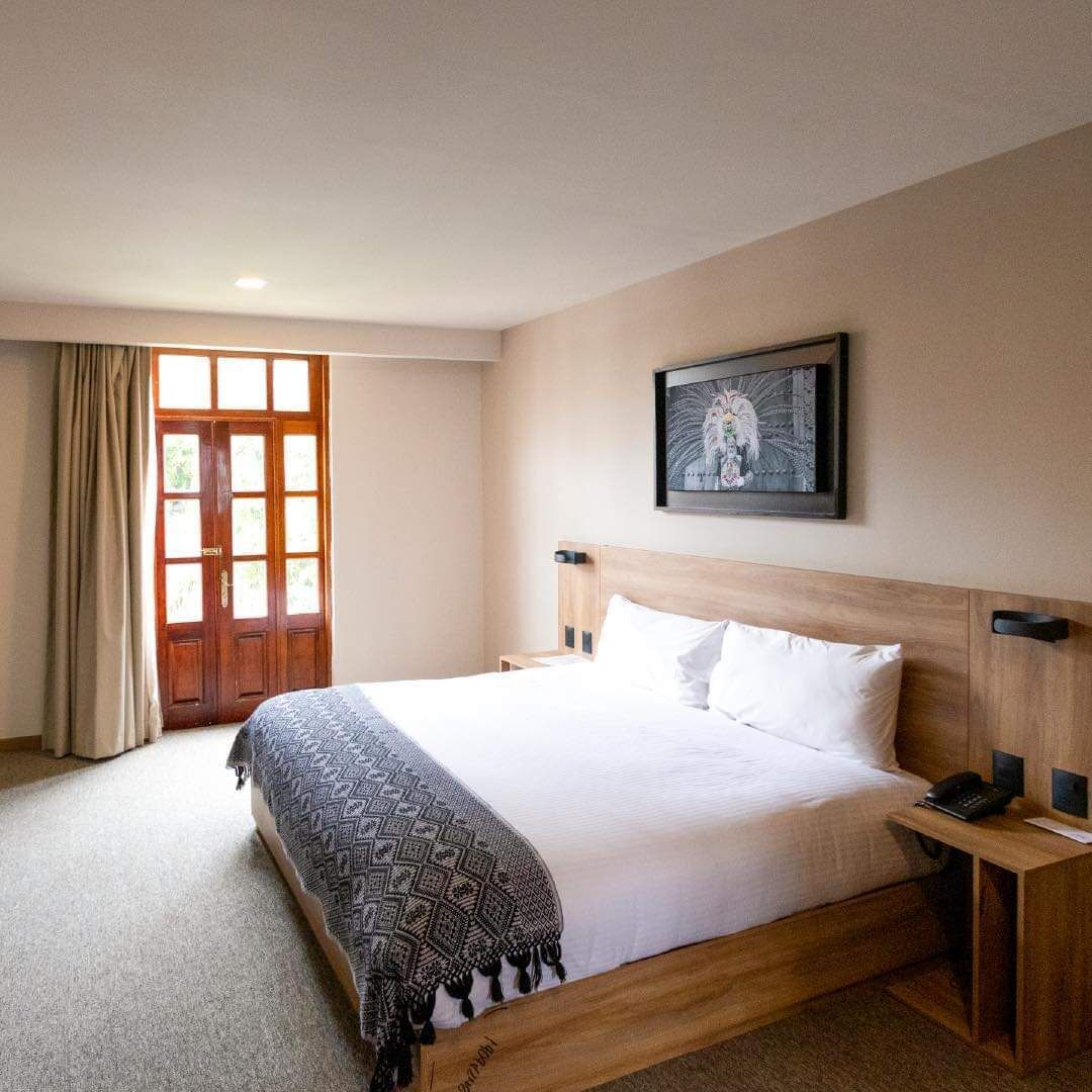 A hotel room with a bed, a bedside table with a phone, a framed picture on the wall, and a door with curtains.