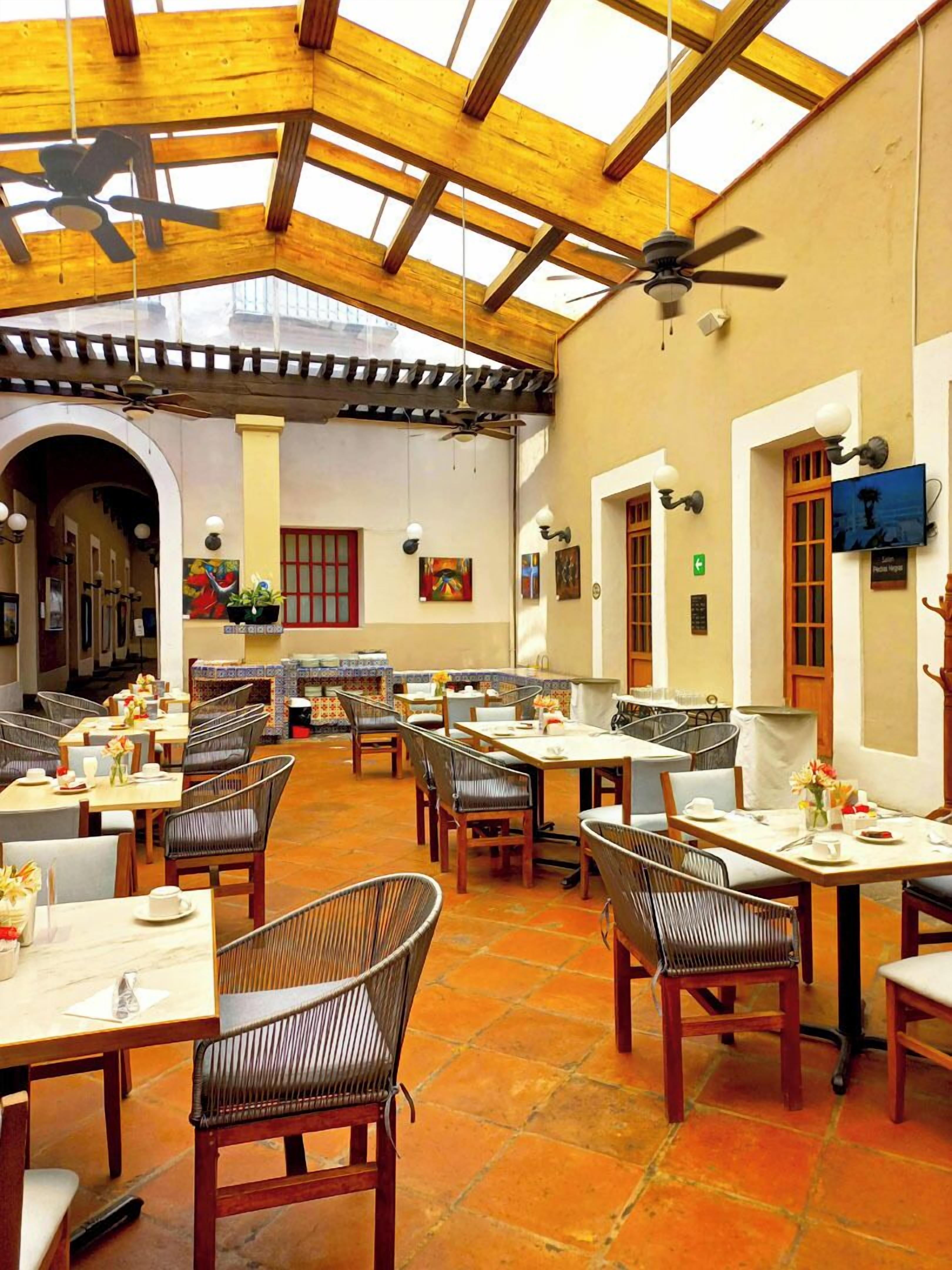 A spacious indoor dining area with wooden tables and chairs, a high ceiling with wooden beams, and a view of the outside through large windows.