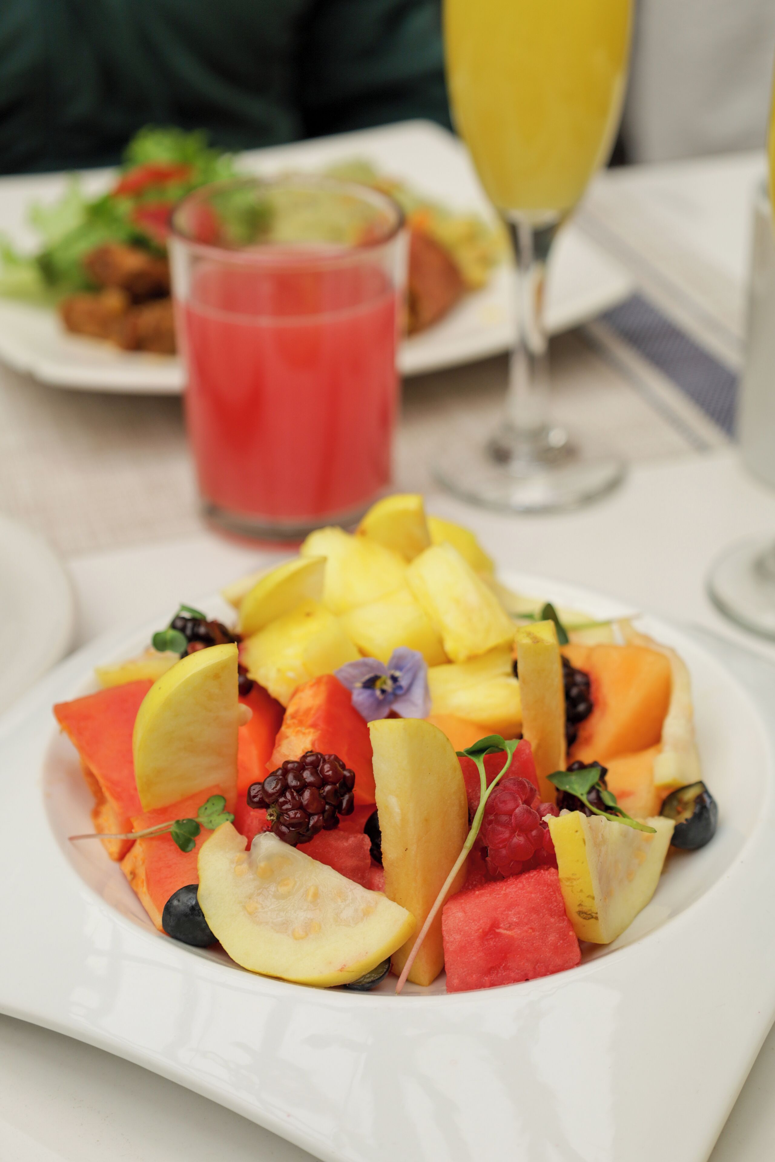 A white plate with a fruit salad, a glass of red drink, and a glass of yellow drink on a table.