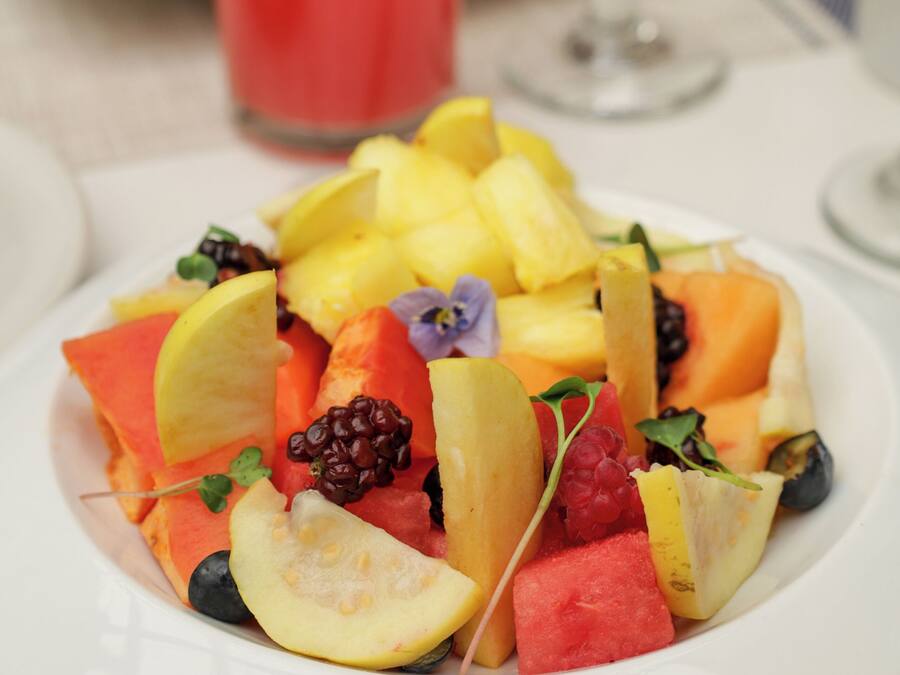 A white plate with a fruit salad, a glass of red drink, and a glass of yellow drink on a table.