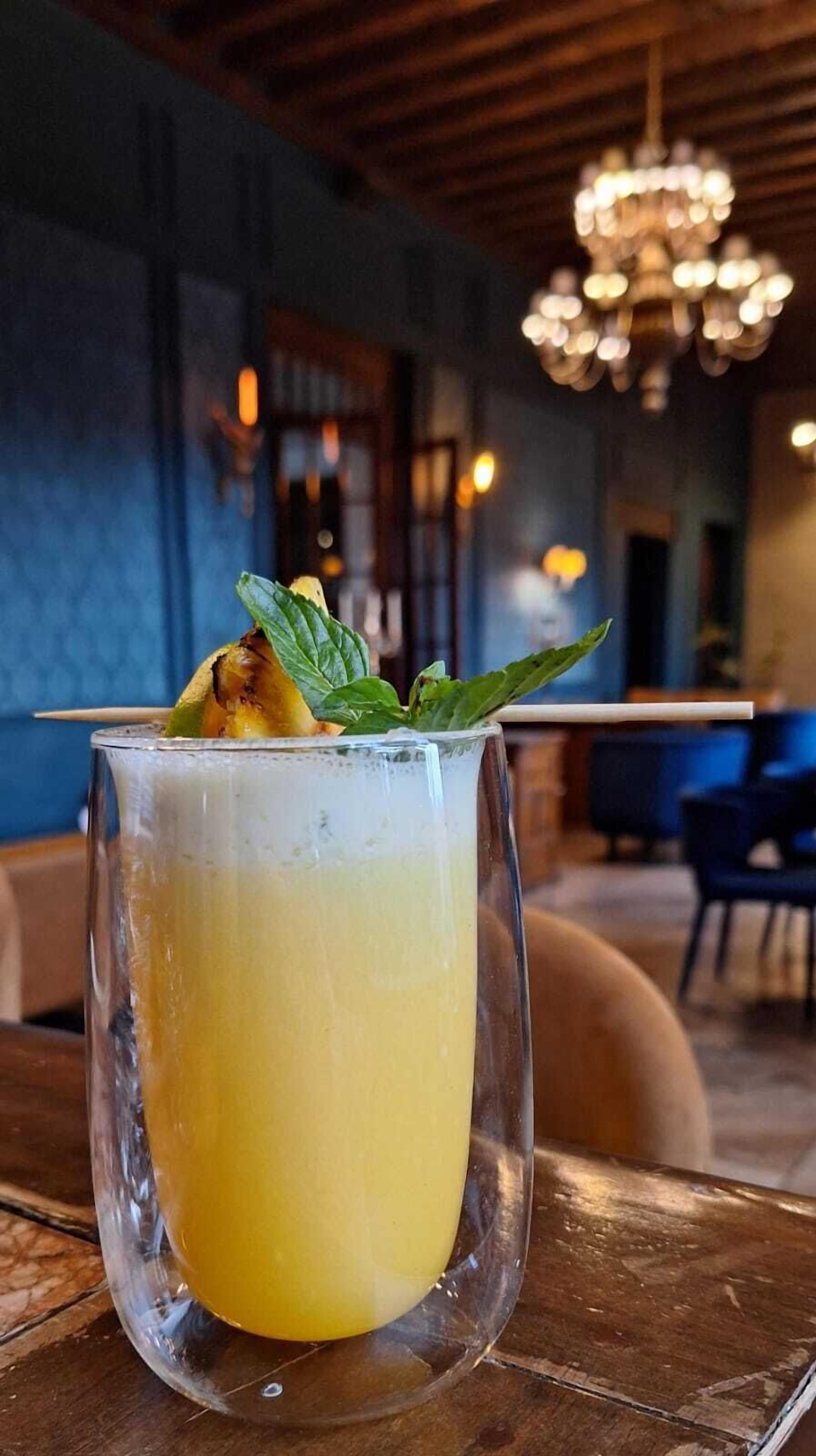 A glass of yellow beverage with a mint garnish on a wooden table in a restaurant with blue walls and a chandelier.