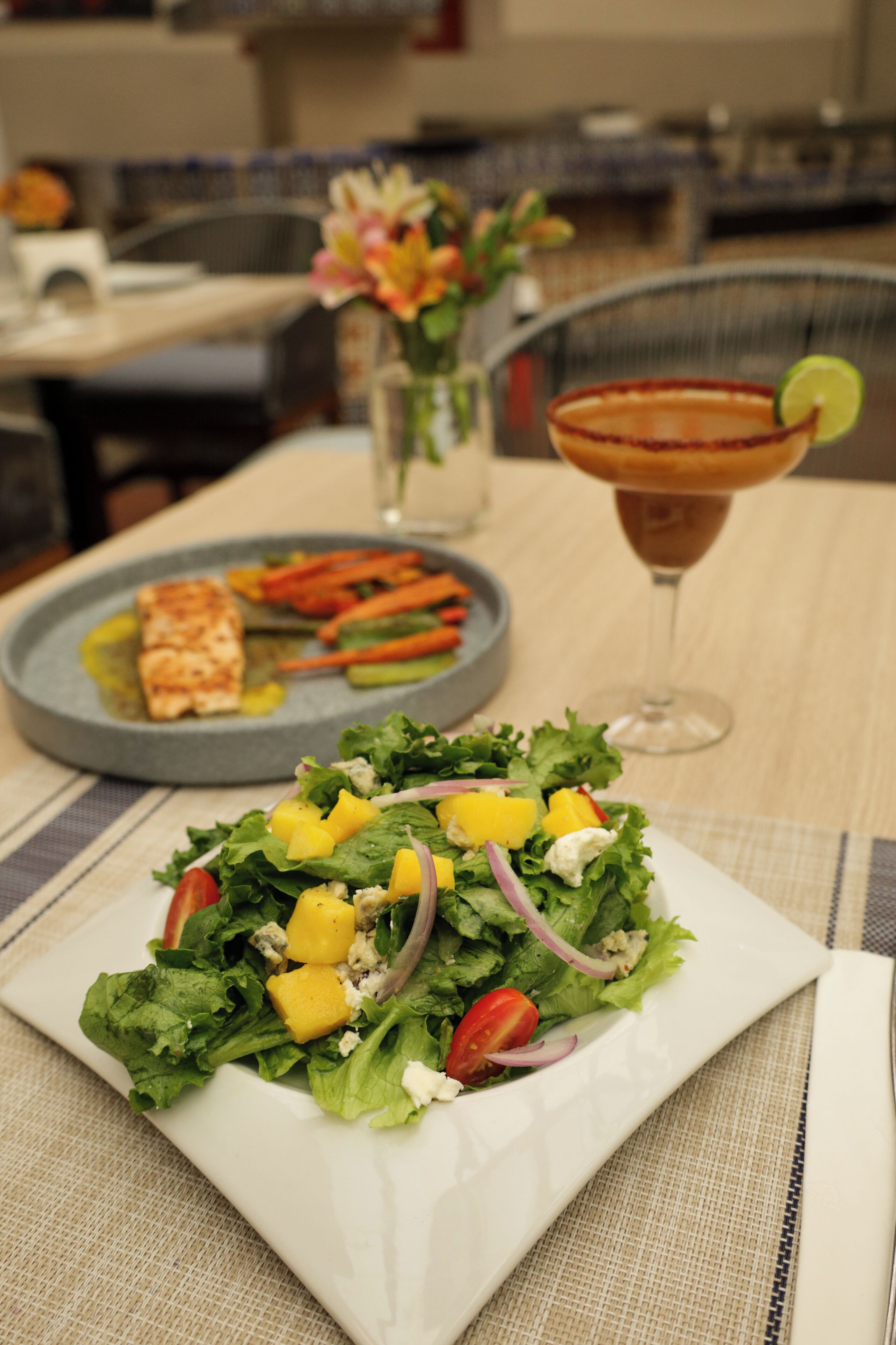A plate of food with a salad and grilled fish, a glass of drink, and a pitcher of flowers on a table.