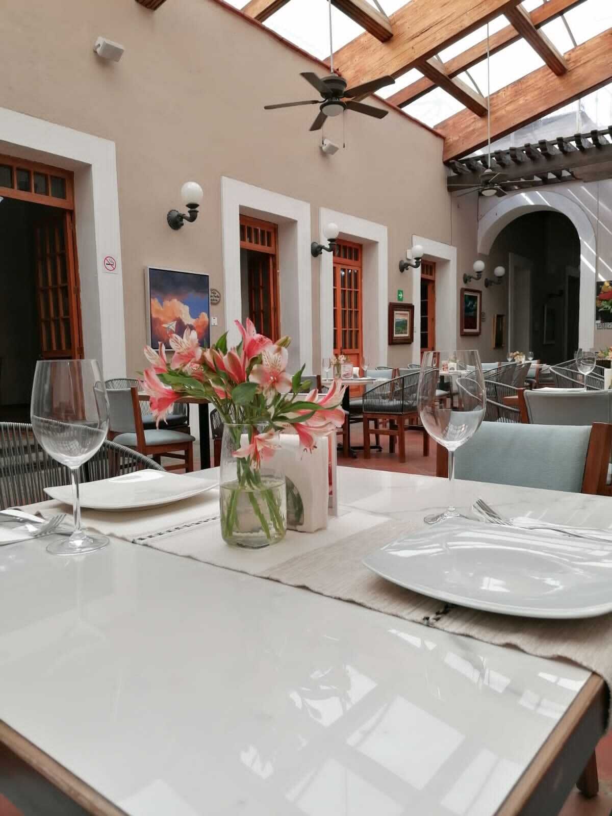 Uma sala de jantar com mesas preparadas para dois, com um vaso de flores cor-de-rosa e taças de vinho.