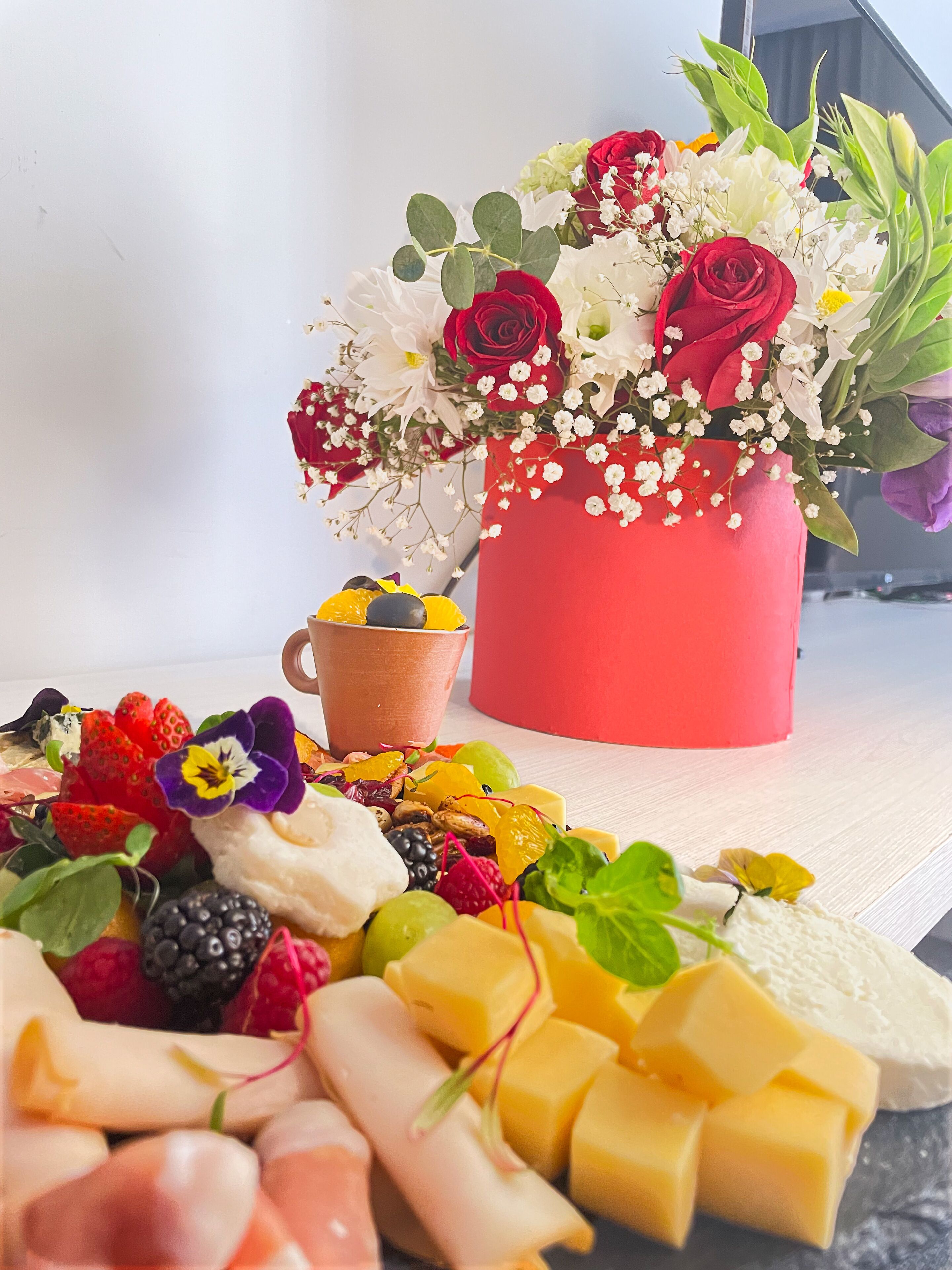 A table with a red vase of flowers, a cup, and an assortment of fruits and cheeses.