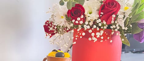 A table with a red vase of flowers, a cup, and an assortment of fruits and cheeses.