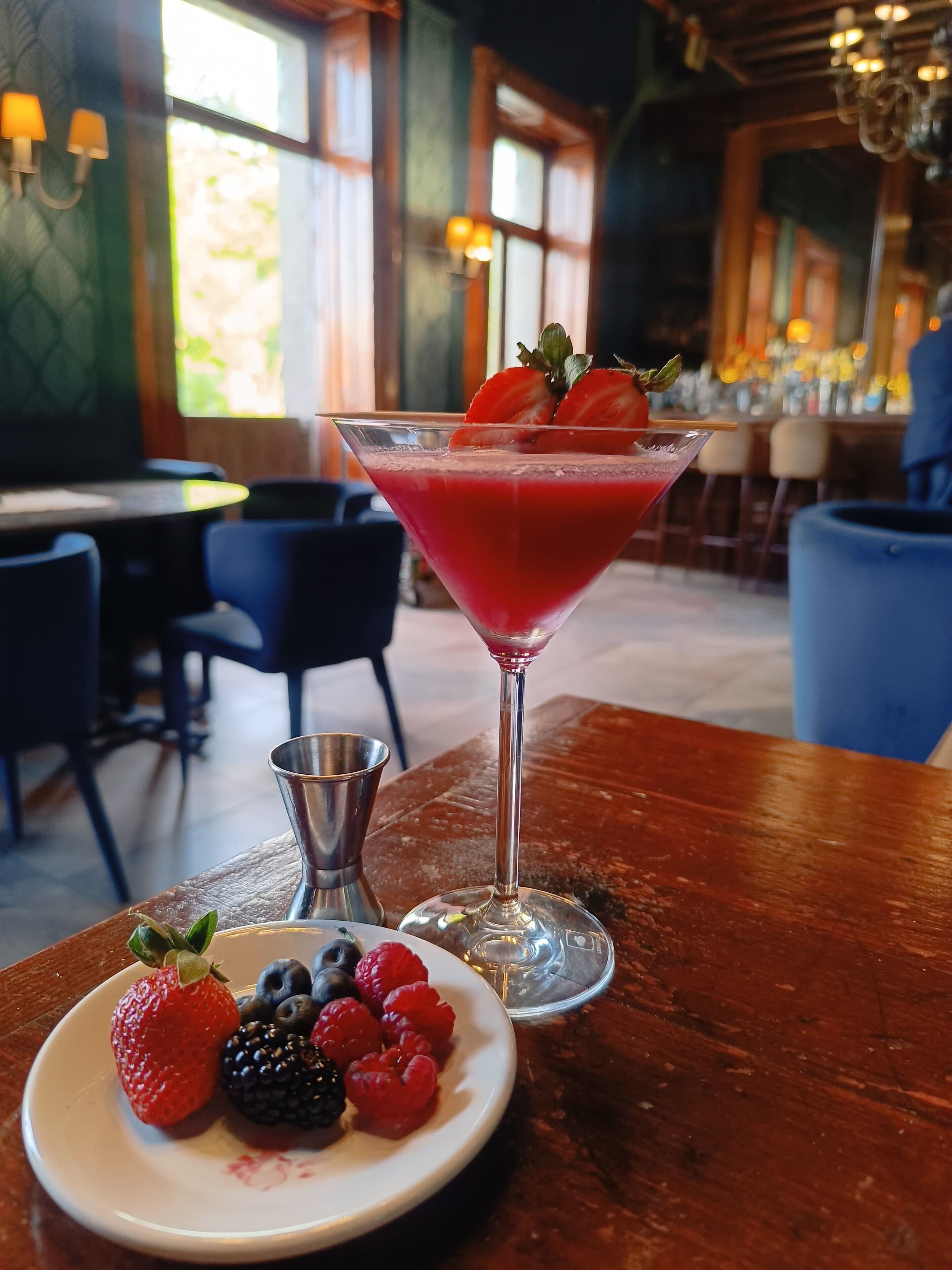 Un bar con una copa de martini que contiene un cóctel rojo adornado con fresas, un plato de bayas variadas y un coctelero sobre una mesa de madera.