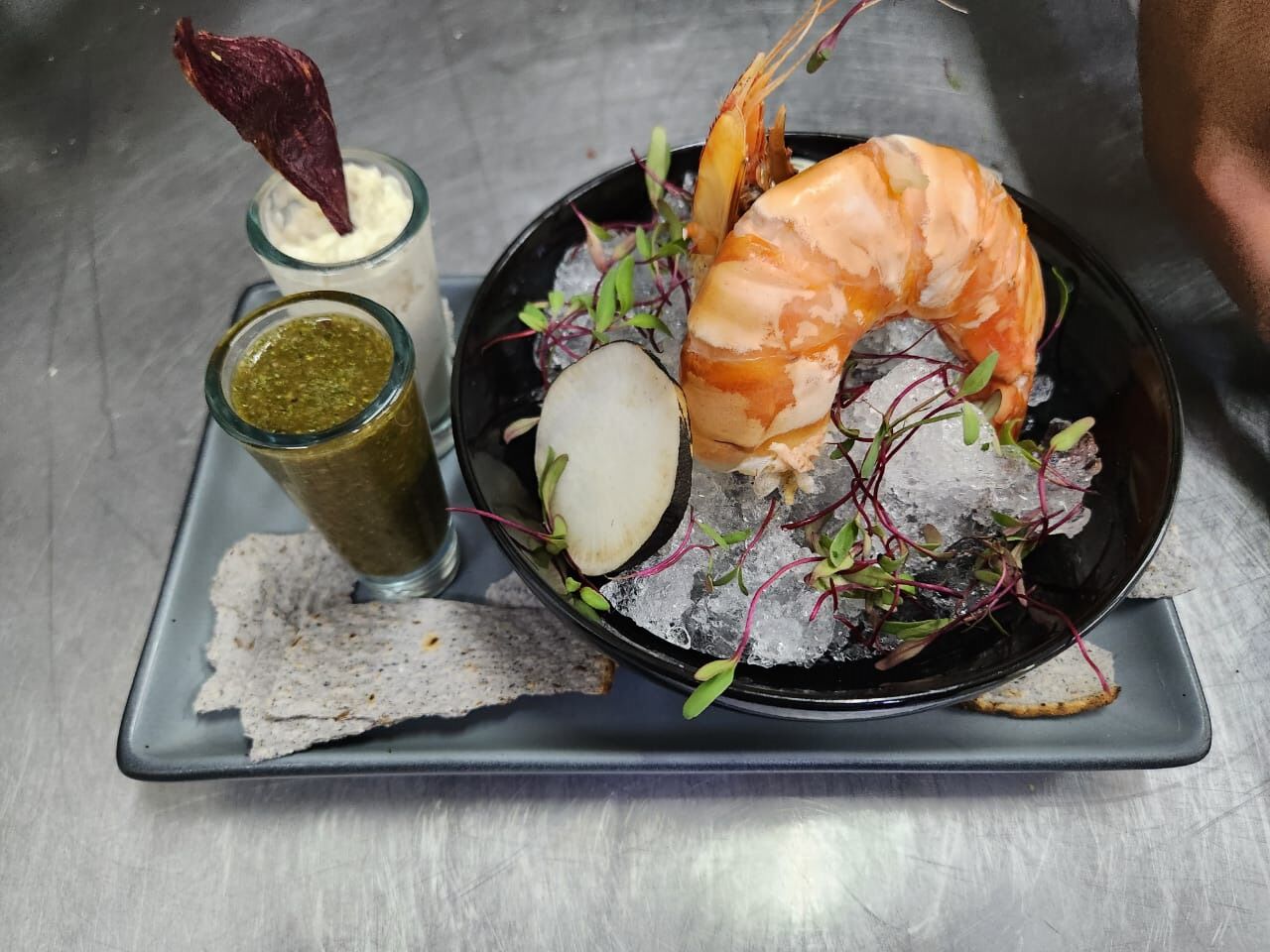 Un plato con un camarón grande, hielo y vegetales verdes, acompañado de un vaso de salsa y una rebanada de pan.