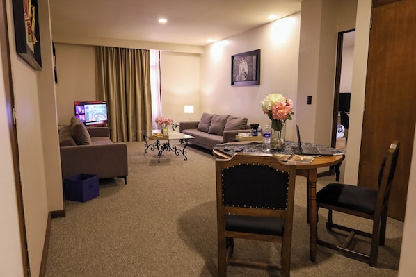 Habitación de hotel con sala de estar que incluye un sofá, una mesa de centro, una computadora portátil y una mesa de comedor con sillas.